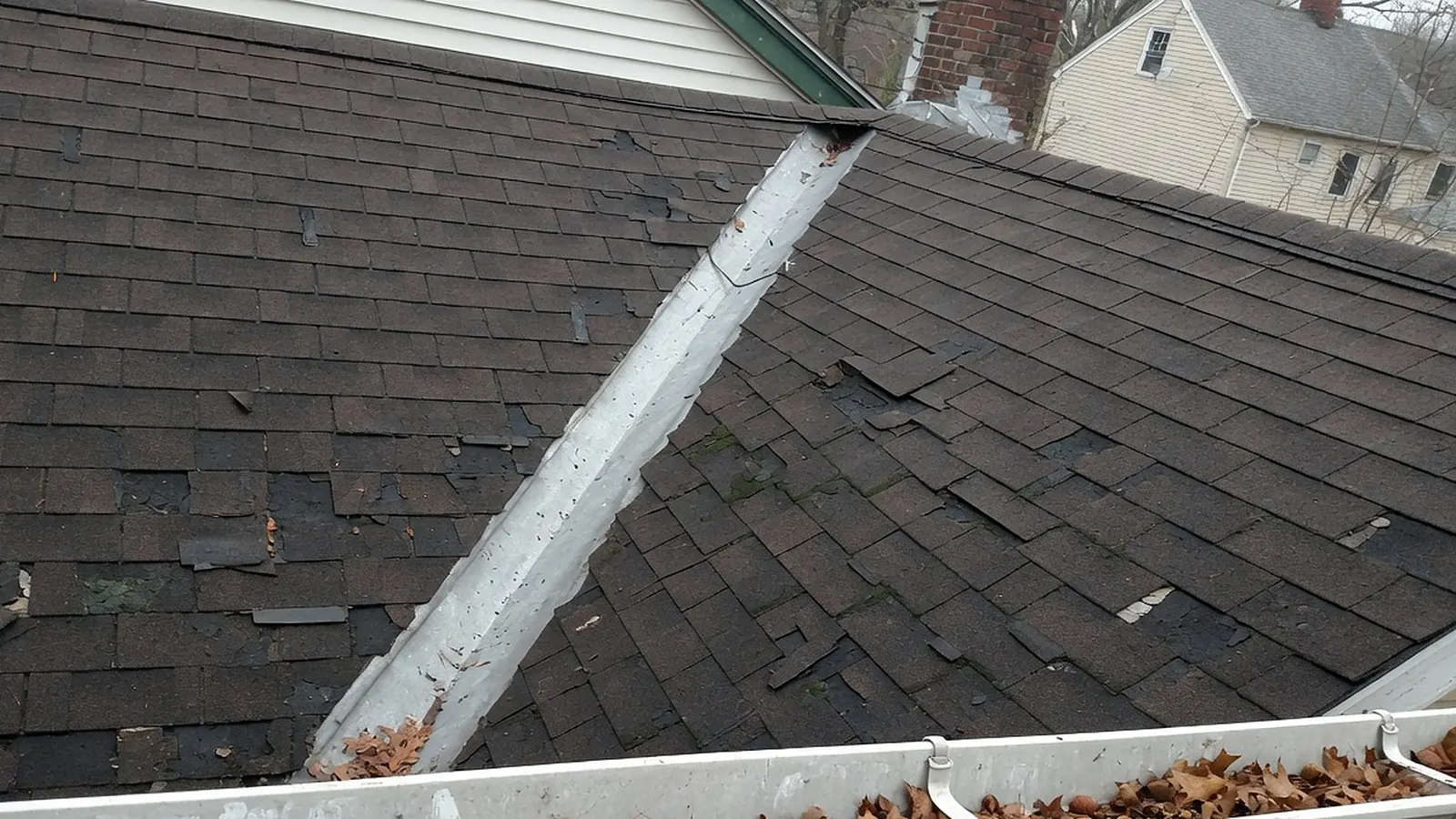 Worn asphalt shingle roof with missing and loose shingles, a patched metal valley and a gutter full of leaves