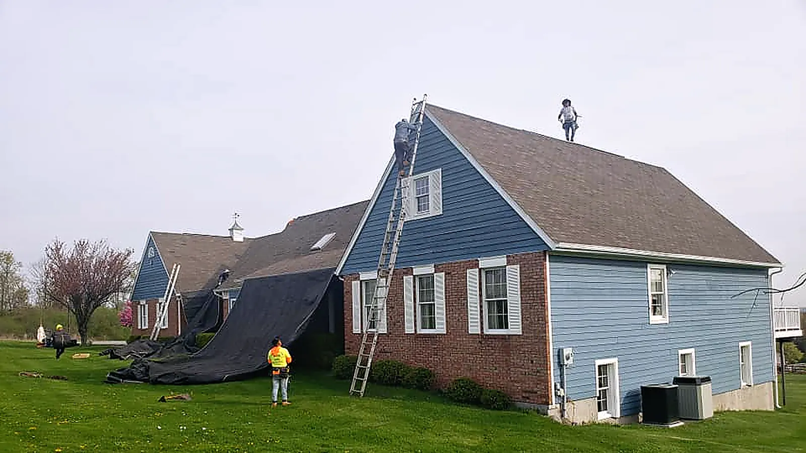 RJ Construction crew tearing off the roof of a blue cape with a brick front, with tarps spread across the lawn