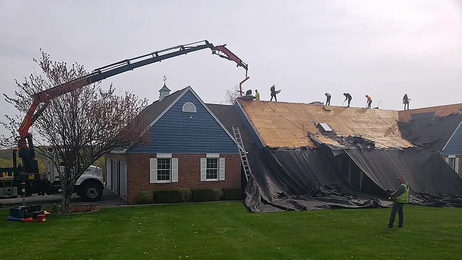 RJ Construction crew on a roof replacement, a boom truck lifting shingles to the roof deck and tarps covering the lawn below